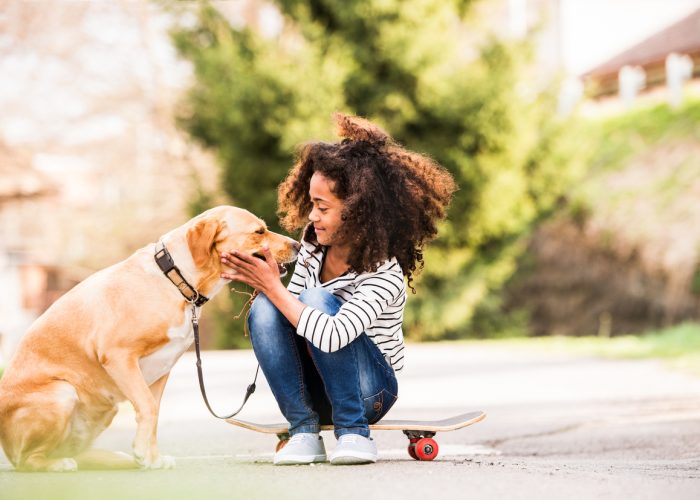 Beautiful african american girl with curly hair outdoors with her cute dog, sitting on skateboard.